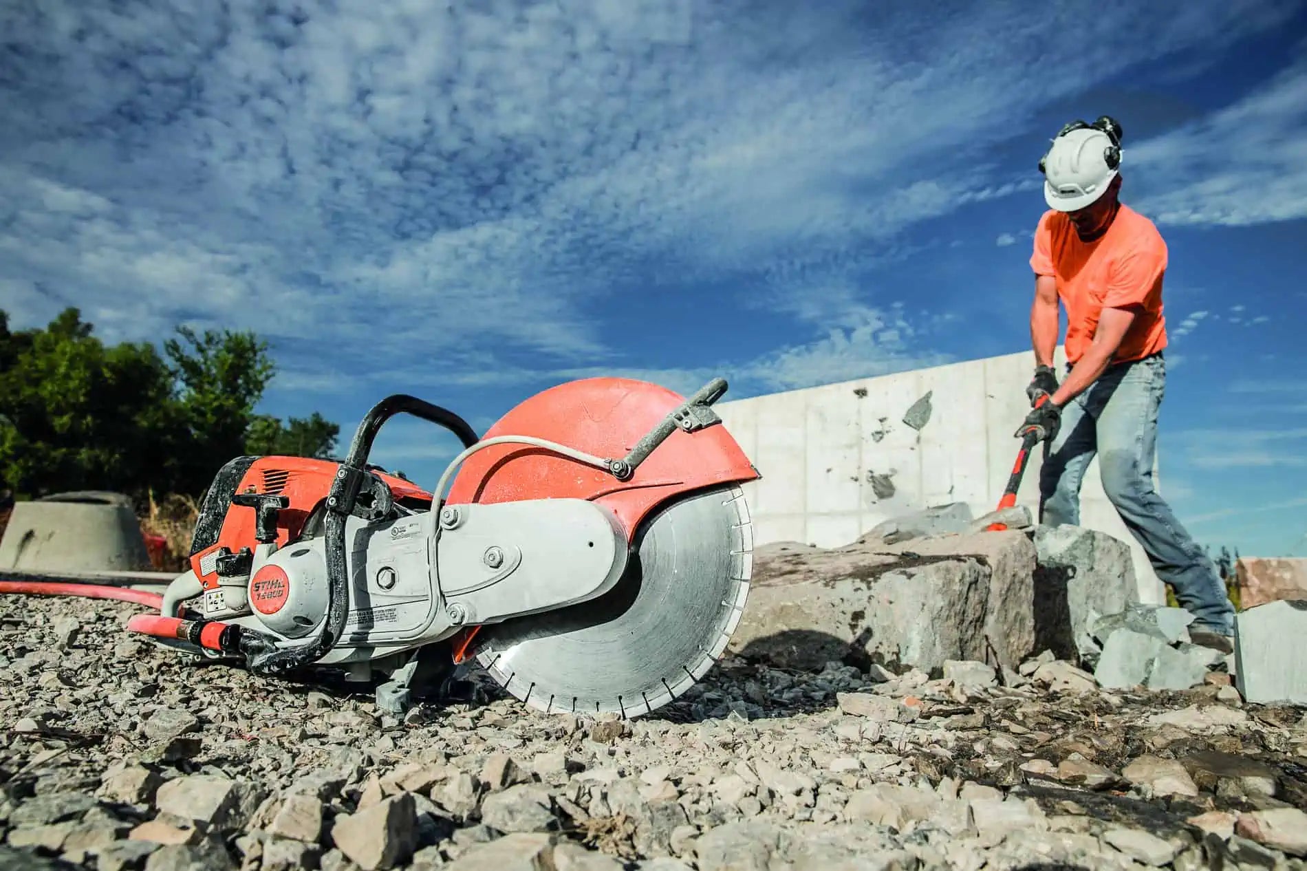 A large red and silver concrete saw with a circular blade lies on a rocky worksite under a blue sky, while a construction worker in an orange shirt and white hard hat uses a hammer to break a large stone nearby.