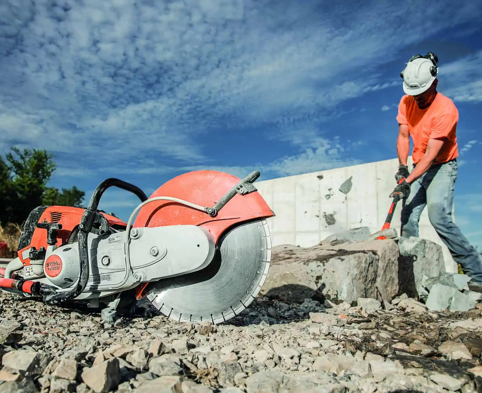 A large red and silver concrete saw with a circular blade lies on a rocky worksite under a blue sky, while a construction worker in an orange shirt and white hard hat uses a hammer to break a large stone nearby.