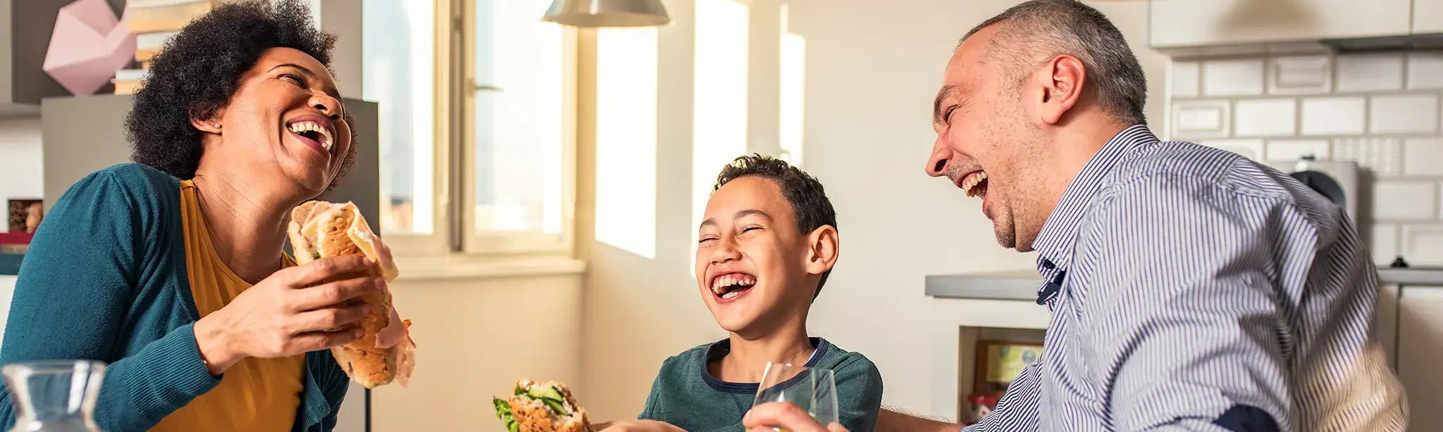 A family of three laughing and eating sandwiches at a dining table in a bright, modern kitchen. The mother on the left wears a teal top over a mustard yellow shirt, the father on the right wears a blue and white striped button-down shirt, and their son in the center wears a dark green t-shirt.