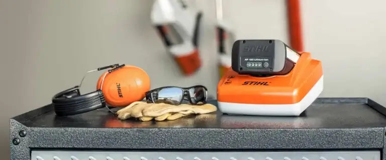 Stihl cordless tools and safety gear arranged on a dark workbench, including an orange-and-white Stihl battery pack, orange earmuffs, black ear protection, beige work gloves, and sunglasses, with a blurred workshop background.