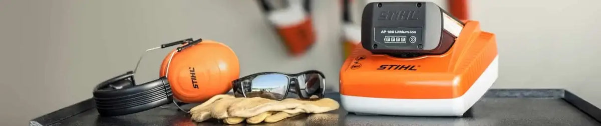 A STIHL cordless tool battery and charger set displayed on a dark tabletop alongside orange ear protection, black cords, sunglasses, and tan gloves. The STIHL charger is bright orange and white, with a black battery resting on top.